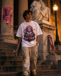 A young man stands on stone steps in front of a classical building, wearing a white graphic t-shirt with a purple and red 'VENI VIDE NON CEDAM' design, beige pants, and holding a skateboard.