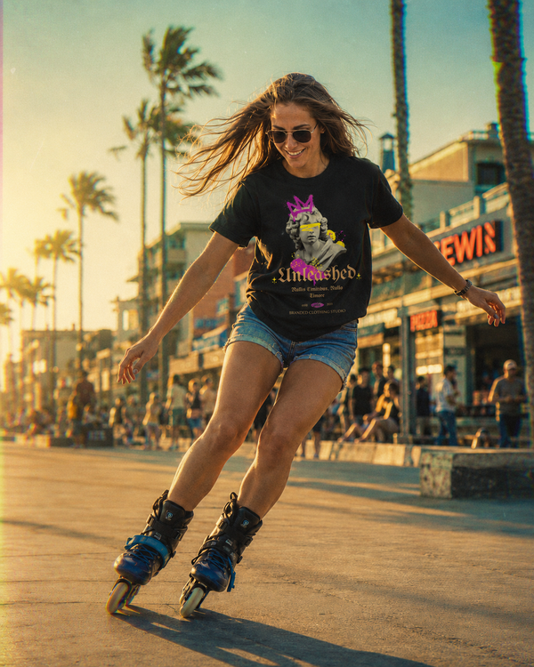 A woman roller skating outdoors on a sunny boardwalk wearing a black graphic t-shirt with a colorful cartoon design, denim shorts, and roller skates. Palm trees and storefronts are visible in the background.